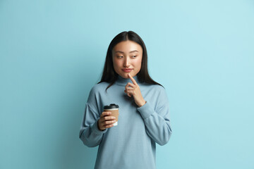 Pondering Woman Holding Coffee to Go. Portrait of Friendly Young Asian Woman Offering Coffee, Holding Drink in Disposable Cup and Smiling. Indoor Studio Shot Isolated on Blue Background 