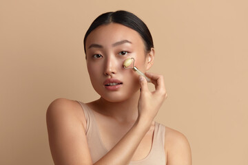 Face massage. Asian Woman Using Jade Facial Roller for Skin Care, Beauty Treatment on Beige Isolate Background. Girl Using Natural Massager Closeup Portrait 