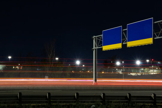 Bright light trails on a highway go under two blank, large directional sign in blue and yellow. 
