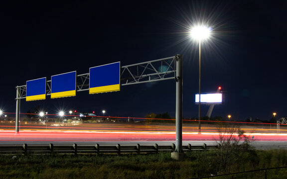 In an urban area, traffic light trails go under three large, blank highway signs at night.