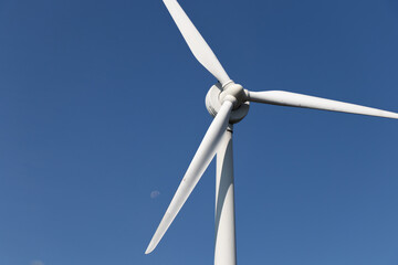 A large wind turbine is seen on a clear, blue sky day with the moon faintly seen in the background.