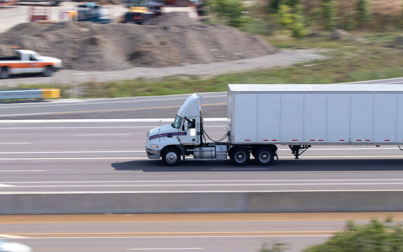 A blank, white semi-truck and trailer is seen on a multi-lane highway on a sunny day.