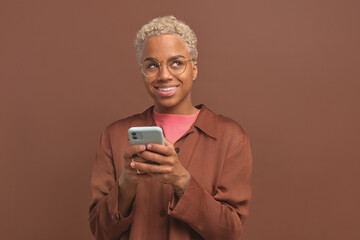 Fototapeta premium Young shy pretty African American woman in glasses with mobile phone in hands dreamily looks up and chats on Internet with new friend from social network stands in brown studio. Smartphone, apps