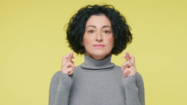 Mature pleading cute woman with wide opened eyes and lifted brows keeping fingers crossed, praying begging about something good, making wish isolated on plain pastel yellow studio background 4K