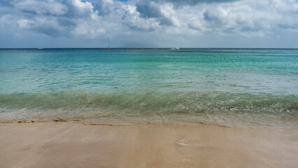 The turquoise wave of the ocean rolls onto the sandy beach. Wet sand glistens. Yachts are visible on the horizon. Clouds in the sky. Seychelles. Mahe