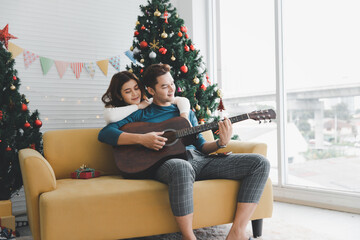 A man playing guitar to woman lover during celebrateing christmas holiday together in living room with christmas tree ornament decoration