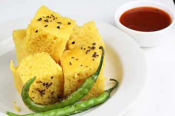 This is a vegetarian culinary dish called Dhokla served in a white plate with chutney in a white bowl on a white background