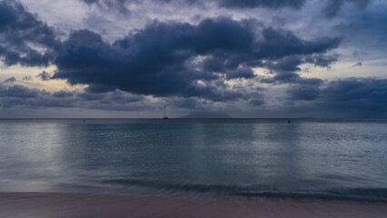 Obraz premium Evening twilight on a tropical island. Blue clouds in the sky. The turquoise ocean is calm. Waves on the sand, a yacht on the horizon. Seychelles. Mahe. Beau Vallon. Long exposure