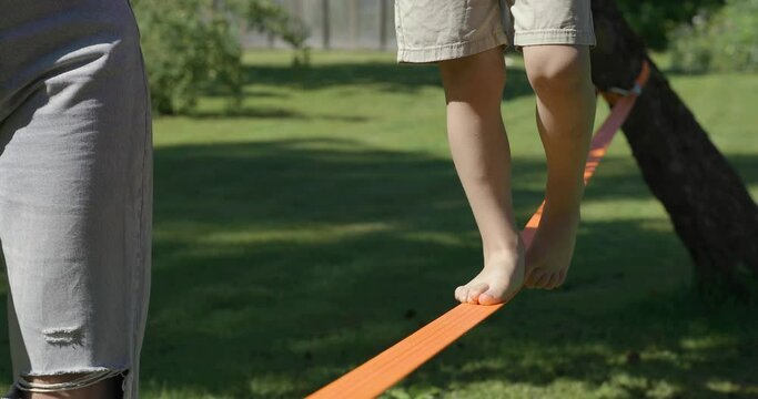 Mother holding kids hand walking on slackline training balance with bear foot on a warm sunny summer day in a green garden. Healthy living concept. Balance and help concept.