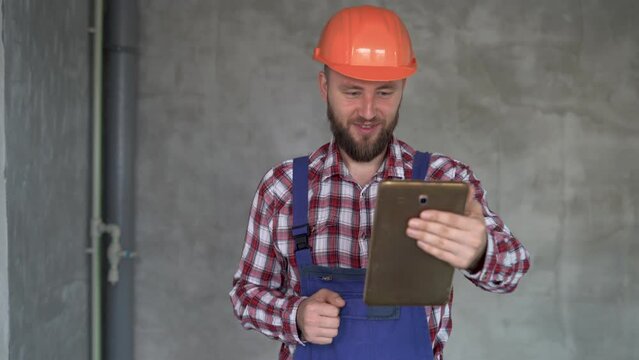 Cheerful bearded builder or foreman has video call while working at construction site, repairman using tablet to check project stage, construction, renovation and technology