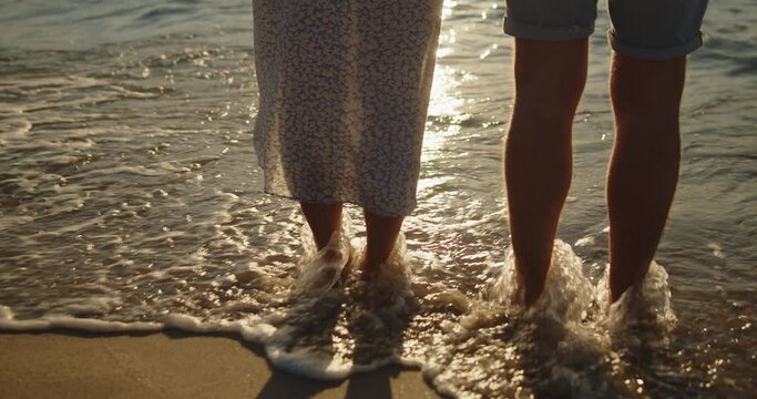 Young loving couple spend time on the beach at sunset. Man and woman stand together on the sea shore. Only legs washed by the waves are taken into the frame. Travel vacation lifestyle.