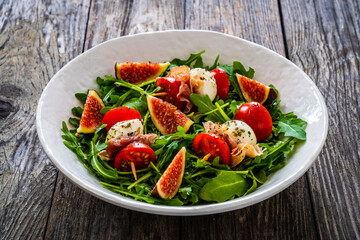 Fresh salad - mozzarella, crudo ham, fig, cherry tomatoes and leafy greens on wooden table
