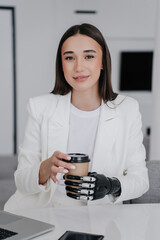 Vertical shot of beautiful brunette girl in white suit with bionic hand prosthesis holding cup of...