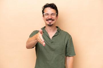 Young caucasian man isolated on beige background shaking hands for closing a good deal