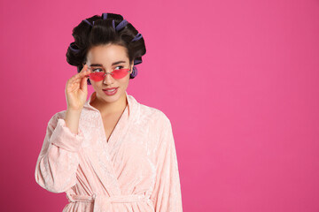 Young woman in bathrobe with hair curlers and sunglasses on pink background, space for text