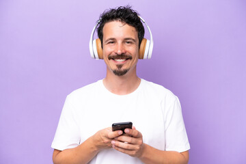 Young caucasian man isolated on purple background listening music with a mobile and looking front