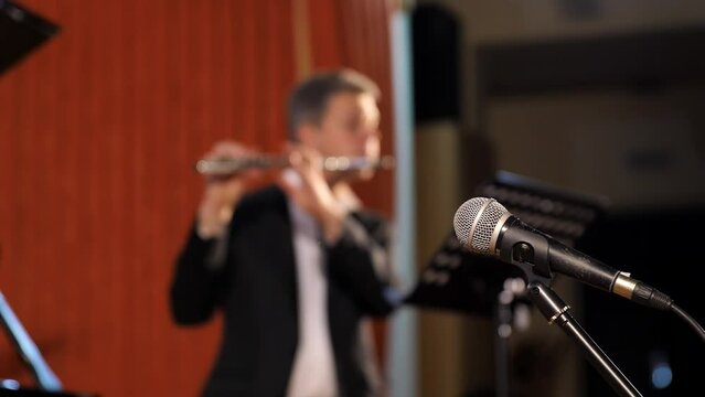Focus on the microphone that stands in the background of a playing flutist during a classical music concert at the Philharmonic. The man plays the flute.