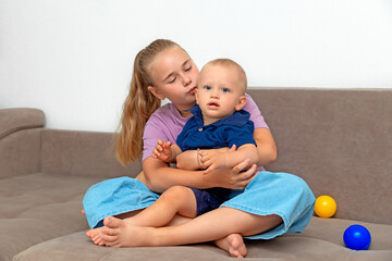 The older sister hugs and kisses her younger brother. The children sitting together on the sofa were laughing and having fun together. Happy childhood