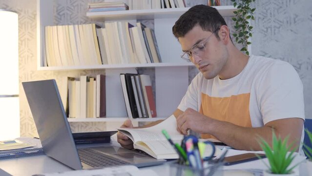 University student reading a book.
The student is reading a book at home. He has laptop and books on his desk.
