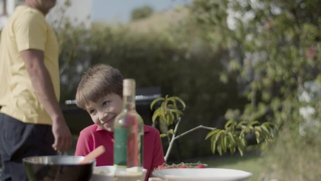 Smiling preteen boy sitting at table in summer garden and smiling, his father walking and smiling in background. Garden party, family leisure concept