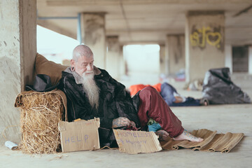 An old homeless man wearing a sweater and blanket sleeps on cardboard looking for help because of...