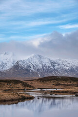 Beautiful Winter panorama landscape image of mountain range and peaks viewed from Loch Ba in Scottish Highlands with dramatic clouds overhead