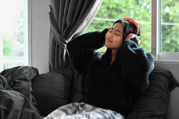 Cheerful young woman listening to music on wireless headphone while sitting in bedroom