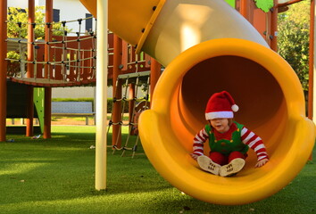 Cute boy in elf costume and Santa Claus hat. The child rides on the children's slide in the playground. Christmas holidays, preschooler. Tropical Christmas
