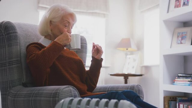 Senior woman sitting in armchair by portable radiator at home with hot drink keeping warm - shot in slow motion