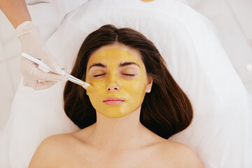 The cosmetologist applies a facial gold mask to the woman's face. Cosmetology and facial skin care in beauty salon. Gold mask. Cosmetic procedure. 