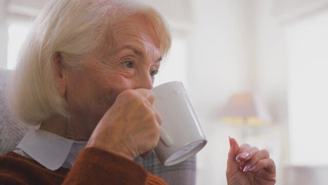 Senior woman sitting in armchair at home with hot drink keeping warm - shot in slow motion