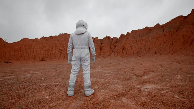 Astronaut in space suit looking at planet crater. Opener from shoulder and rear view of standing man in space suit on far red surface planet.