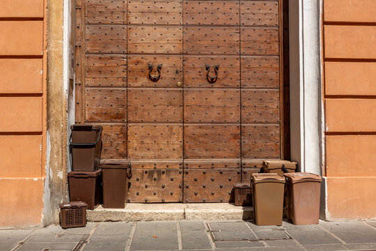 Brown plastic bins with biological waste on the sidewalk in front of a historic old wooden door in the village of Foligno in the central Italian region of Umbria in the province of Perugia.