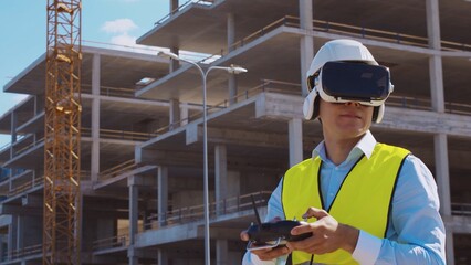 Professional drone operator in virtual reality helmet standing in front of construction site. Builder holding remote controller. Office building and crane background.