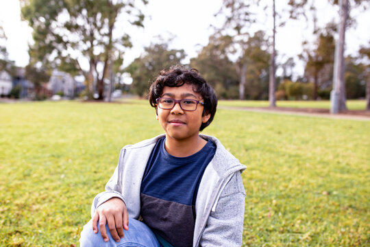 smiling boy with curly hair and eyeglasses wearing grey hoodie with blue undershirt on a field