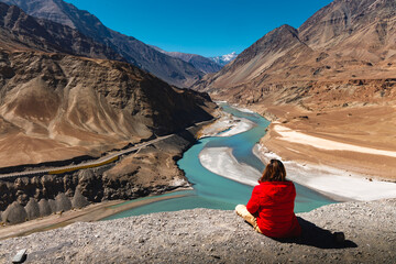 Man in red jacket sit on cliff and look at Sangam or confluence of Indus and Zanskar Rivers at Leh, Ladakh, India.