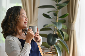 Old woman with a cup of coffee sit in the living room and looked outside.
