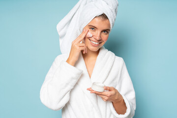 Woman applying facial cream on cheek dressed in bathrobe with head wrapped in towel