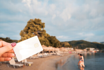 White Bank Card In Woman Hand On Background Of Hotel Beach, Family Comes Out Of Sea, Moraitika, Corfu, Greece. The Concept Of Payment For Vacation. Copy Space.