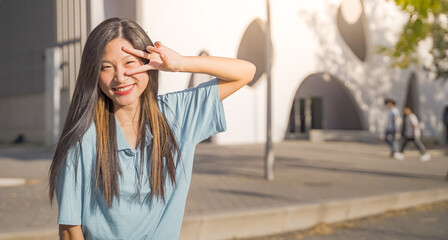 Fototapeta premium Portrait of a beautiful asian woman smiling and looking at camera victory fingers outdoors in the student campus banner copy space.