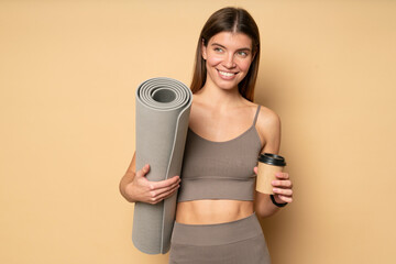 Fit woman in sport top holding rolled mat and cup of coffee after yoga class on brown background