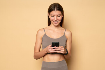 Portrait of fit woman standing on brown background with phone watching online workout tutorial