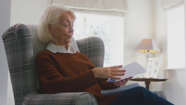 Worried senior woman at home sitting in armchair looking at energy bill during cost of living energy crisis - shot in slow motion