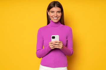 Smiling young woman holding phone, isolated on yellow background