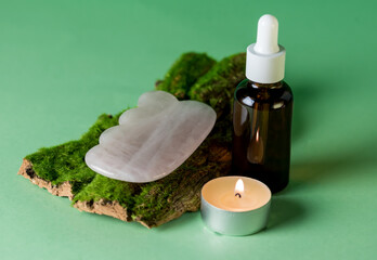 Fototapeta na wymiar Glass Bottle with Oii Gua Sha Stone for Face Massage on Tree Bark on Green Background Burning Candle Cosmetic Facial Skin Care and Spa Natural