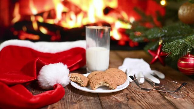 bitten oatmeal cookie and an unfinished glass of milk on a wooden table against the background of Santa Claus clothes, a Christmas tree and a fireplace