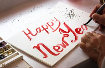 Artist draws inscription Happy New Year with red paint, brush on white sheet of paper. Hand made DIY postcard for Christmas 2023. Festive background, sequins in the form of stars. Painter creates art.