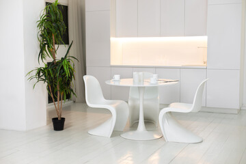 Stylish white dining room with large round table and designer chairs, cups coffee, large houseplant in pot. White plastic chairs pantone. Modern Kitchen interior design with furniture.	