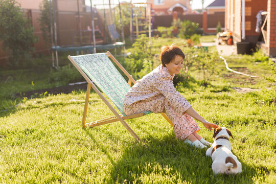 Young woman in pajama resting in chair on green lawn on sunny summer day and pet jack russell terrier dog - village and country life