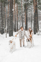 Fototapeta na wymiar Love romantic young couple girl, guy in snowy cold winter forest walking with pet, dog of hunting breed russian borzoi. Sighthound, wolfhound owner. Having fun, laughing. Stylish fur coat, woolen hat